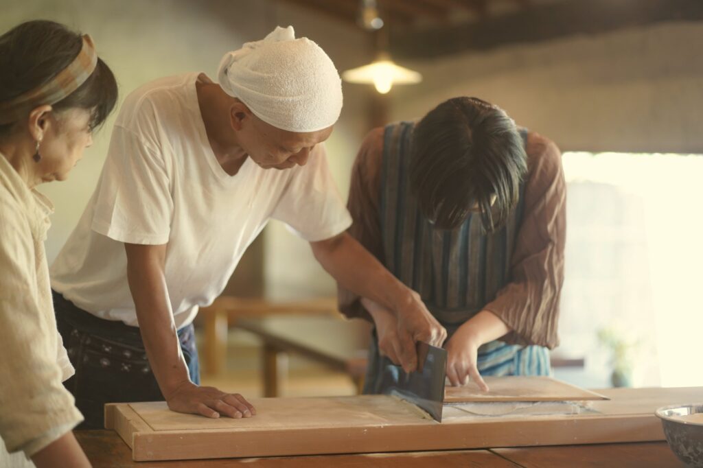 そば職人に手ほどきを受けながら、包丁でそばを切る体験参加者