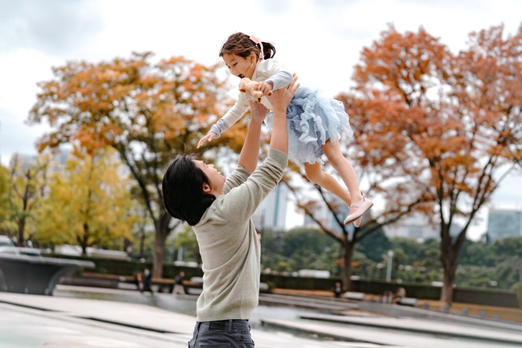 皇居外苑の紅葉を背に、娘を高く抱き上げて笑う父親